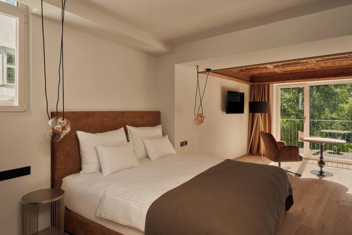 A bedroom with a large bed, a round side table, a chair, a TV, and a balcony with a table and chair.