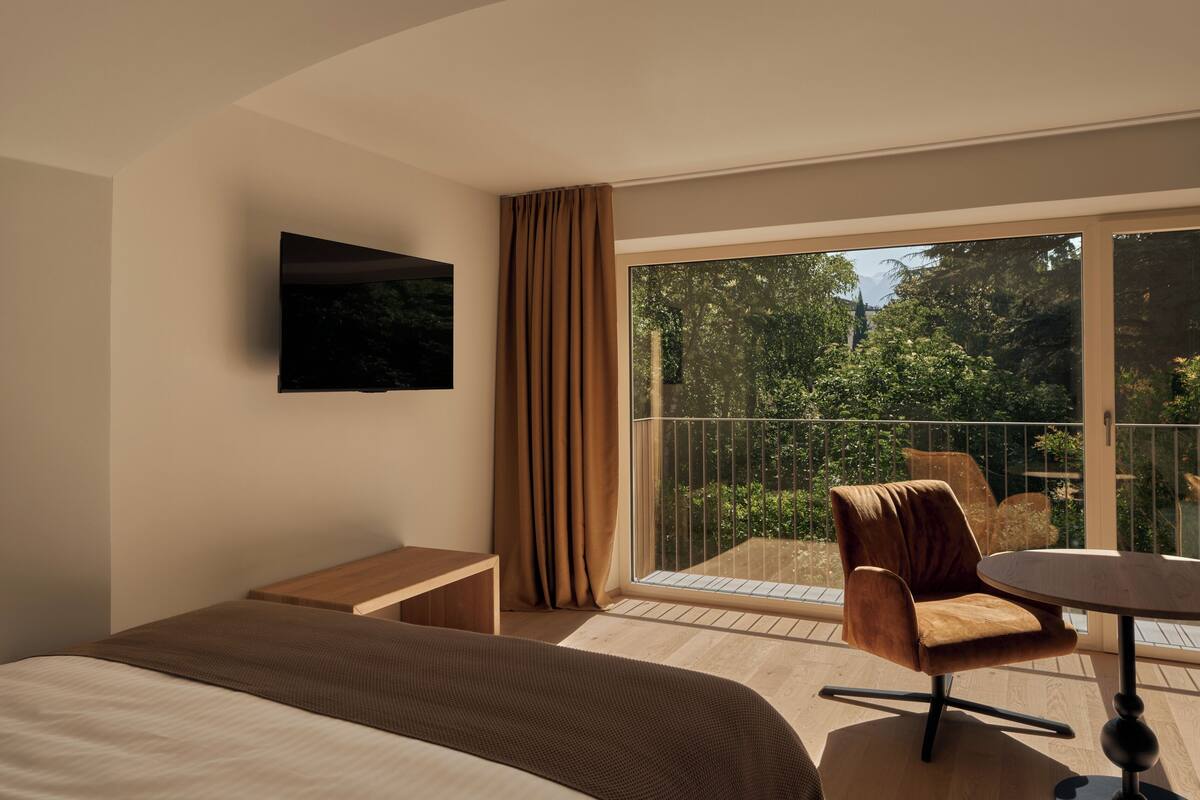 A hotel room with a bed, a small table, a chair, a TV mounted on the wall, and a balcony with a view of greenery.