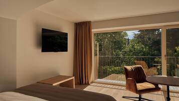 Habitación de hotel con cama, una mesita, una silla, televisor en la pared y balcón con vistas a la vegetación.