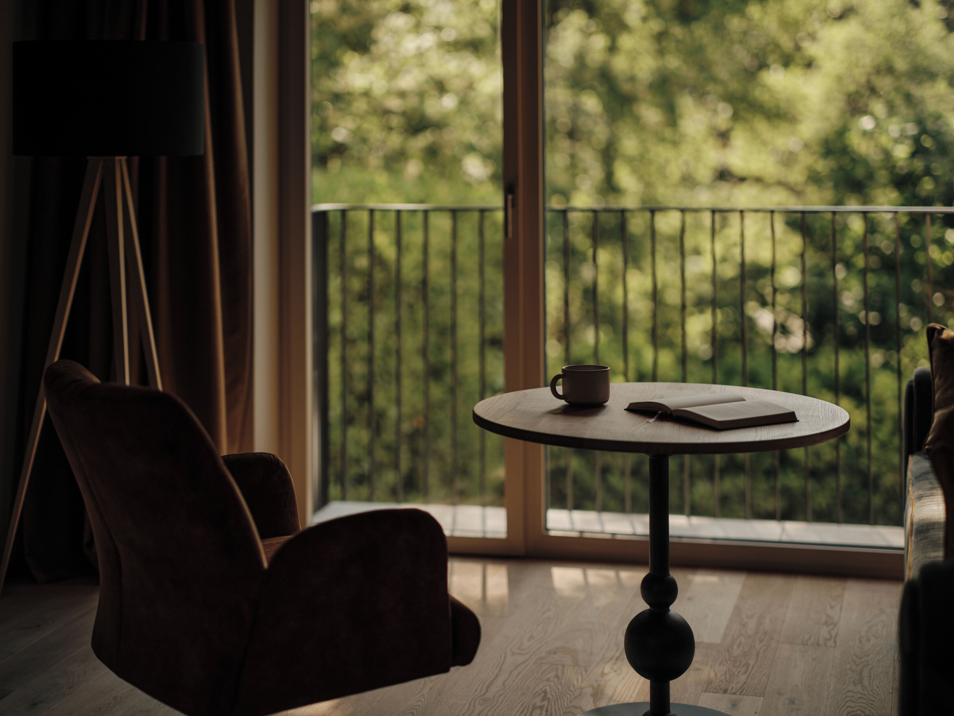 Ein Zimmer mit rundem Tisch, einem Stuhl, einem Fenster mit Blick auf Bäume und einem Balkongeländer.