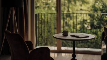 Ein Zimmer mit rundem Tisch, einem Stuhl, einem Fenster mit Blick auf Bäume und einem Balkongeländer.