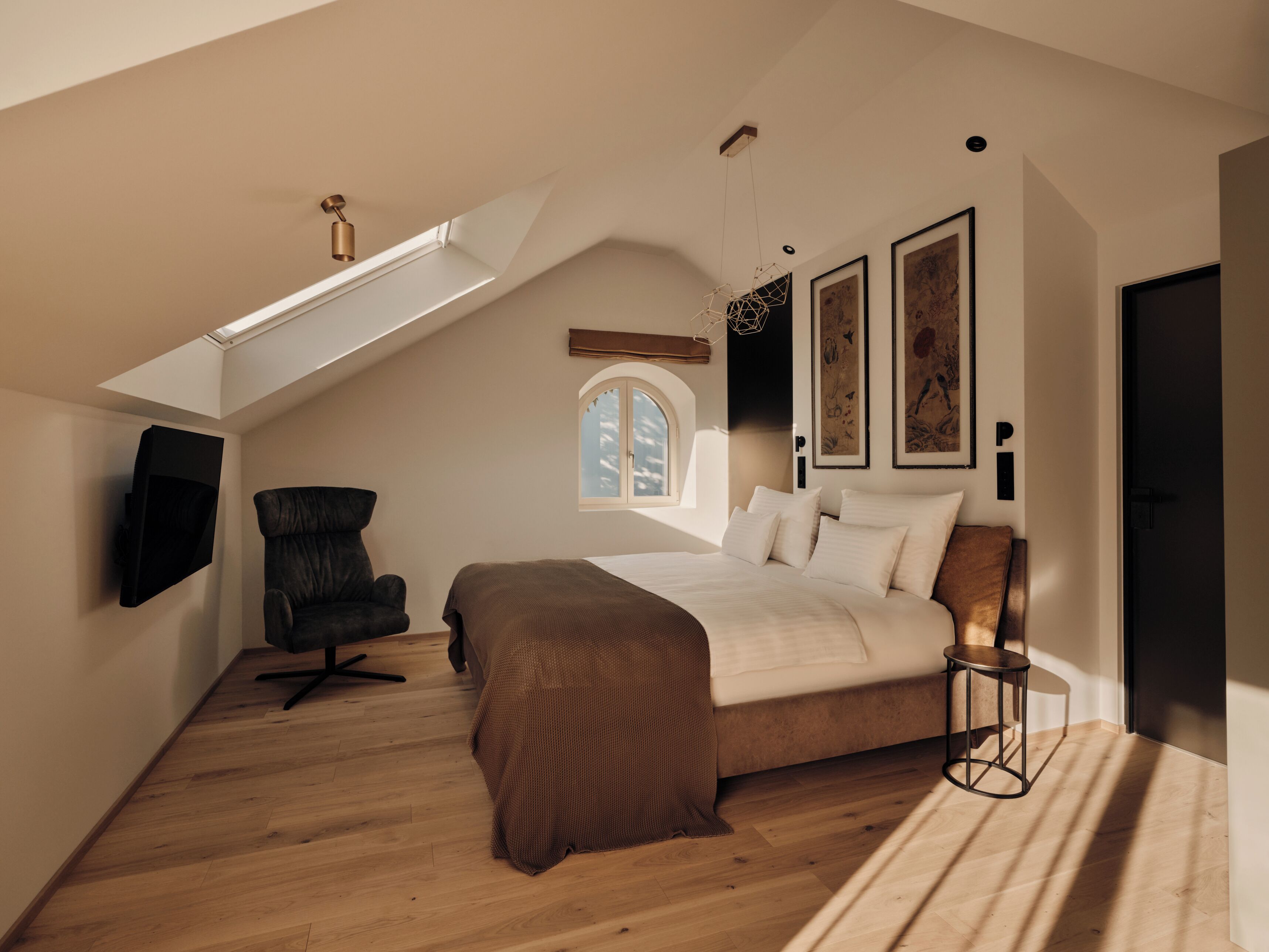 Ein Schlafzimmer mit schrägem Dach, einem Bett, einem Sessel, einem an der Wand befestigten Fernseher und Kunstwerken.