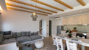 A modern kitchen with wooden beams, a dining area with white chairs, and a sofa with patterned cushions.