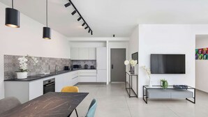 Una cocina moderna con armarios blancos, encimera negra y salpicadero de azulejos. Hay un comedor con una mesa de mármol y sillas, y un televisor montado en la pared.