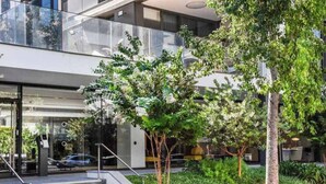 Un edificio moderno con entrada de cristal, escaleras exteriores y un jardín ajardinado con árboles.