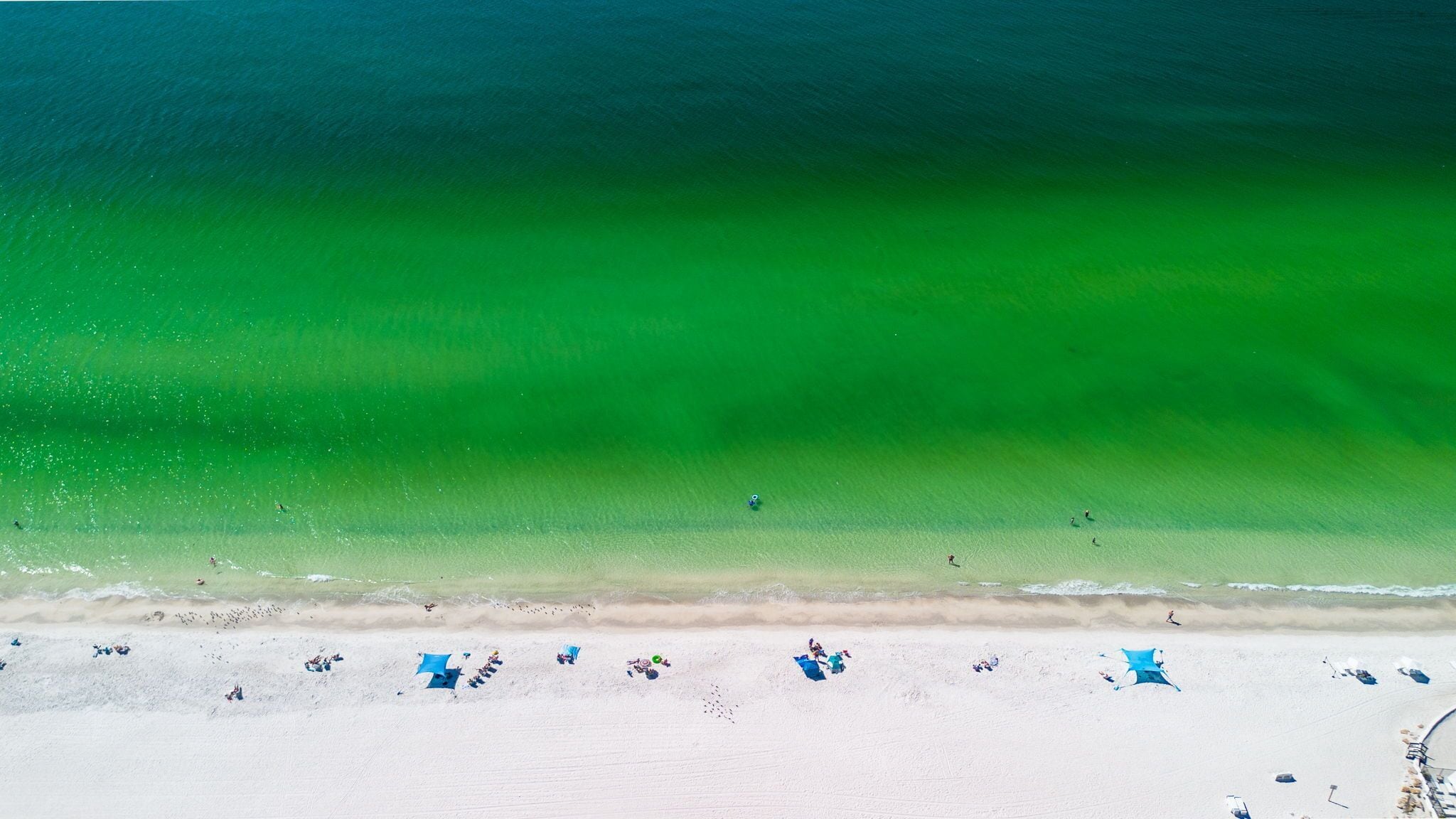 An aerial view of a beach with clear turquoise water, white sand, and several blue beach umbrellas.
