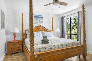 Uma cama com dossel e cabeceira de madeira, mesa de cabeceira com gavetas, ventilador de teto e janela com cortinas.