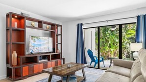 A living room with a TV, wooden entertainment unit, sofa, and a view of greenery through a glass door. - Endless Memories - 1801   “Anna Maria Island” (Bradenton Beach)