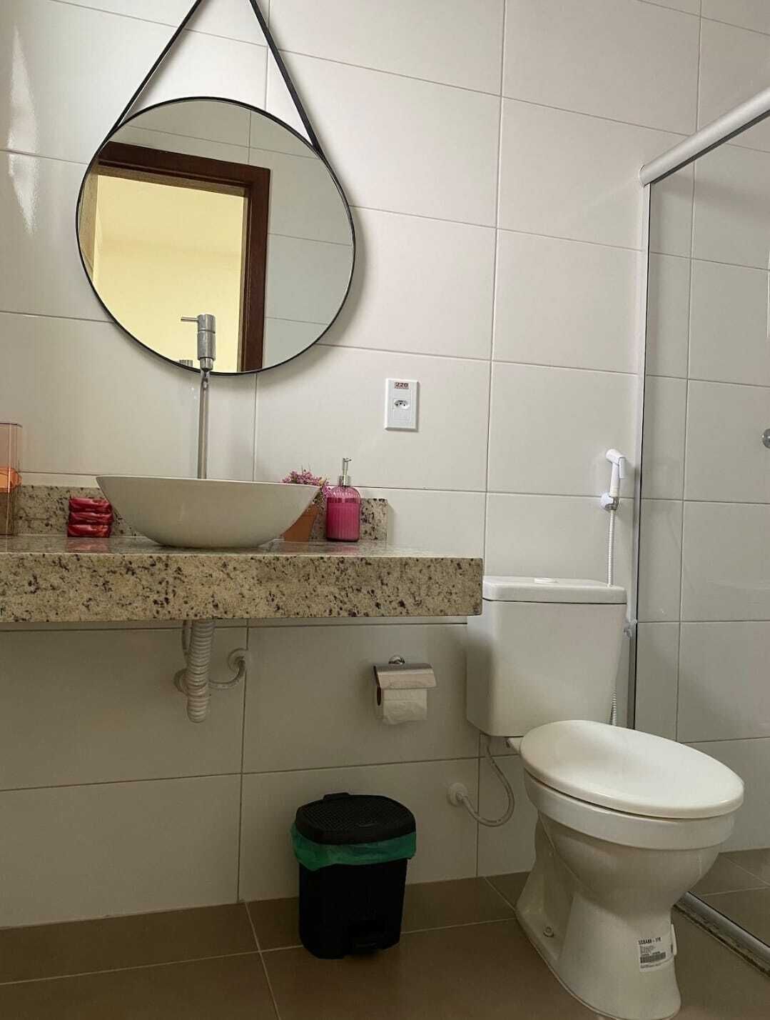 A bathroom with a round mirror, a sink with a vessel, a toilet, and a trash bin.