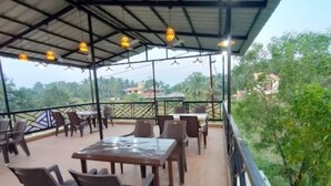 An outdoor dining area with tables and chairs, a metal roof, and a view of greenery and buildings in the background. - Beach Aura (Nagaon)