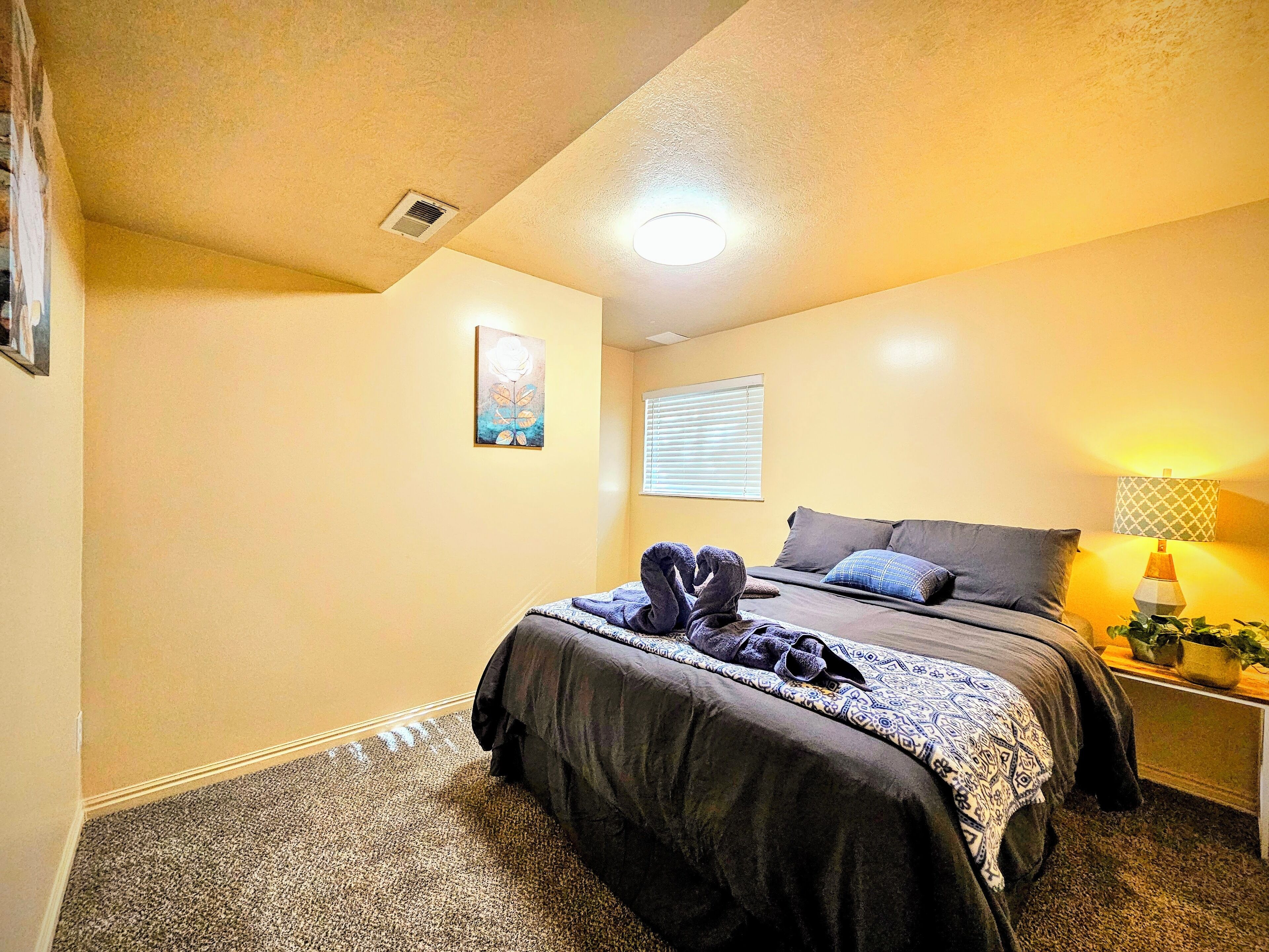 A bedroom with two beds, a ceiling light, a window with blinds, and a nightstand with a lamp and a plant.