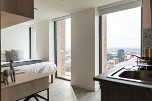 A modern bedroom with a large window, a city view, a bed with a gray and white duvet, and a wooden desk with a giraffe figurine. - Student Only Martha Street (Glasgow)