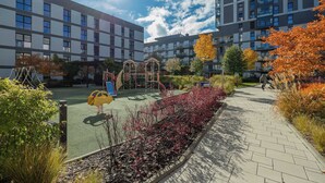 A playground area with a climbing structure, surrounded by a landscaped garden with red foliage, next to a multi-story residential building with balconies.