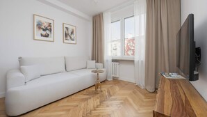Un salón moderno con un sofá blanco, una mesita redonda y una mesa de centro de madera. Hay dos cuadros enmarcados en la pared, una ventana con cortinas y un televisor de pantalla plana montado en la pared.