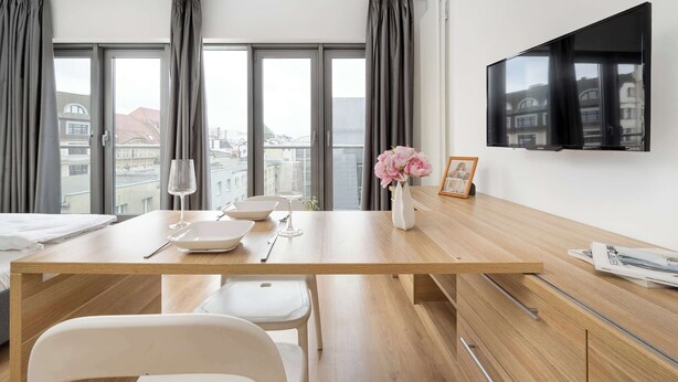 A modern hotel room with a wooden dining table, white chairs, a flat-screen TV mounted on the wall, and large windows with curtains. - Stylish Studio in the Center | Air Conditioning | Parking (Wrocław)