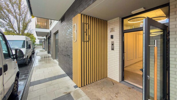 A hotel entrance with a modern design, featuring a wooden panel wall, a glass door, and a tiled pathway leading to parked vehicles.