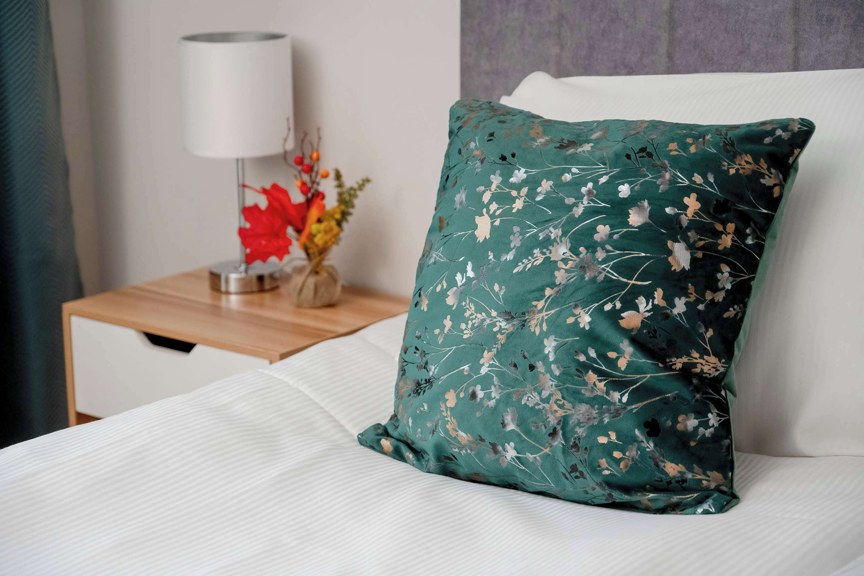 A neatly made bed with a green floral pillow and a white pillow, a bedside table with a lamp and a vase of flowers.