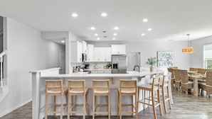 A modern kitchen with white cabinetry, a central island with bar stools, and a dining area with wicker chairs. - 30a Tropical Breeze Beach Home Near Beach! 6Seater Lsv, Pool, Gated Community (Santa Rosa Beach)