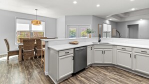 A modern kitchen with white cabinets, a dishwasher, and a dining area with wicker chairs and a wooden table. - 30a Tropical Breeze Beach Home Near Beach! 6Seater Lsv, Pool, Gated Community (Santa Rosa Beach)
