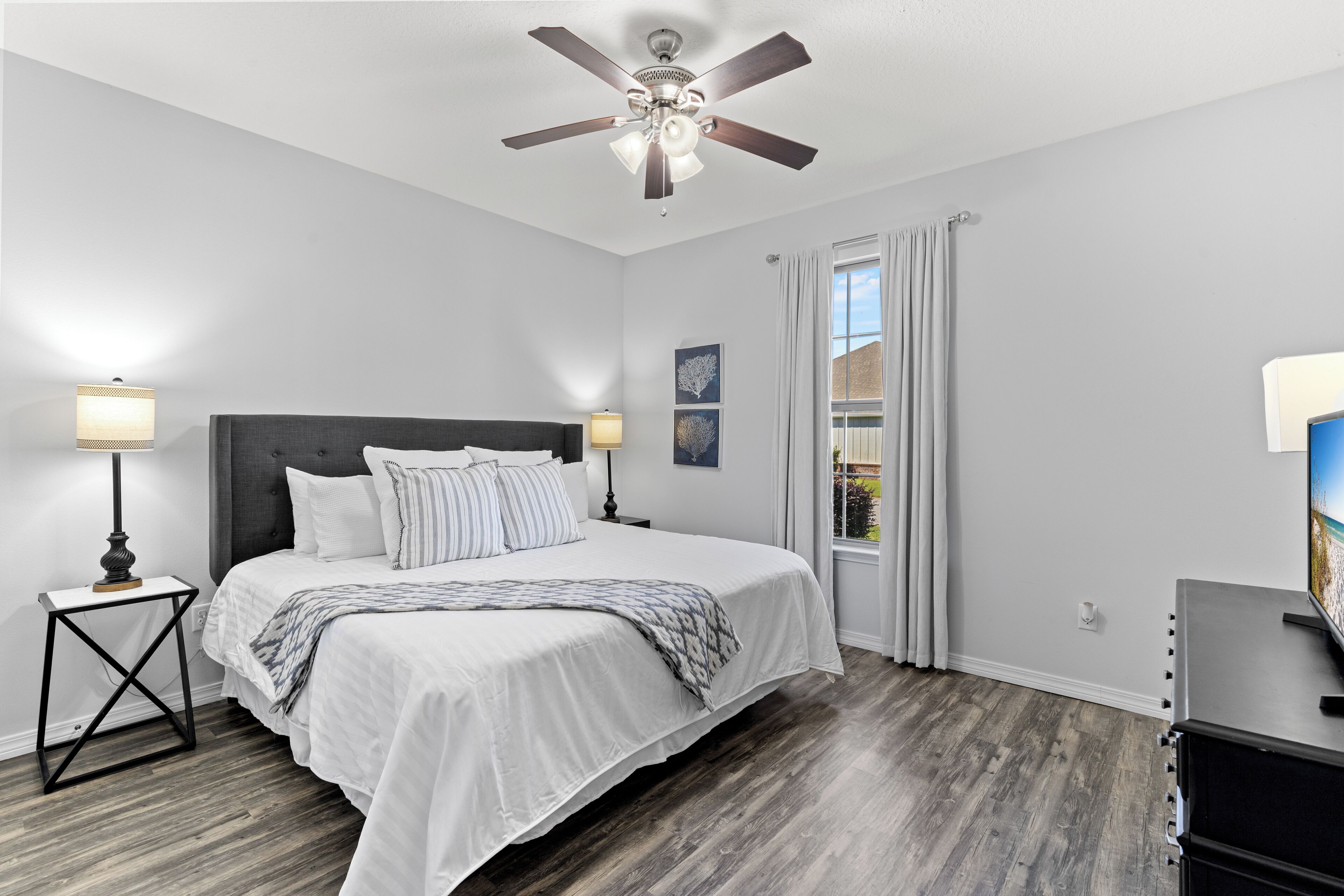 A bedroom with a bed, two bedside tables with lamps, a ceiling fan, and a window with curtains.