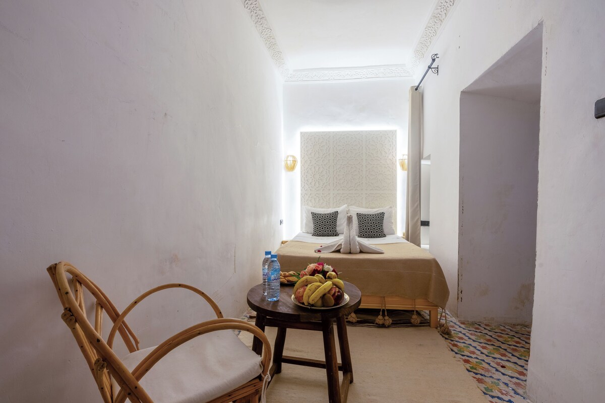 A small, cozy room with a bed, a table with a plate of fruit, a chair, and a bottle of water.