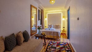 Une chambre d’hôtel avec un lit, un canapé, un miroir, une petite table avec un vase et des bananes, et un tapis coloré.