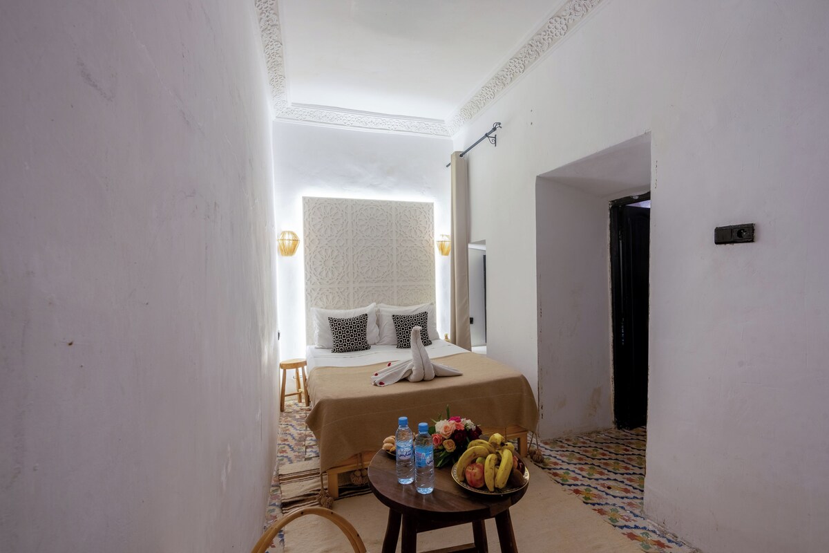 Quarto de hotel com cama, uma pequena mesa com garrafas de água e frutas, e um piso estampado.