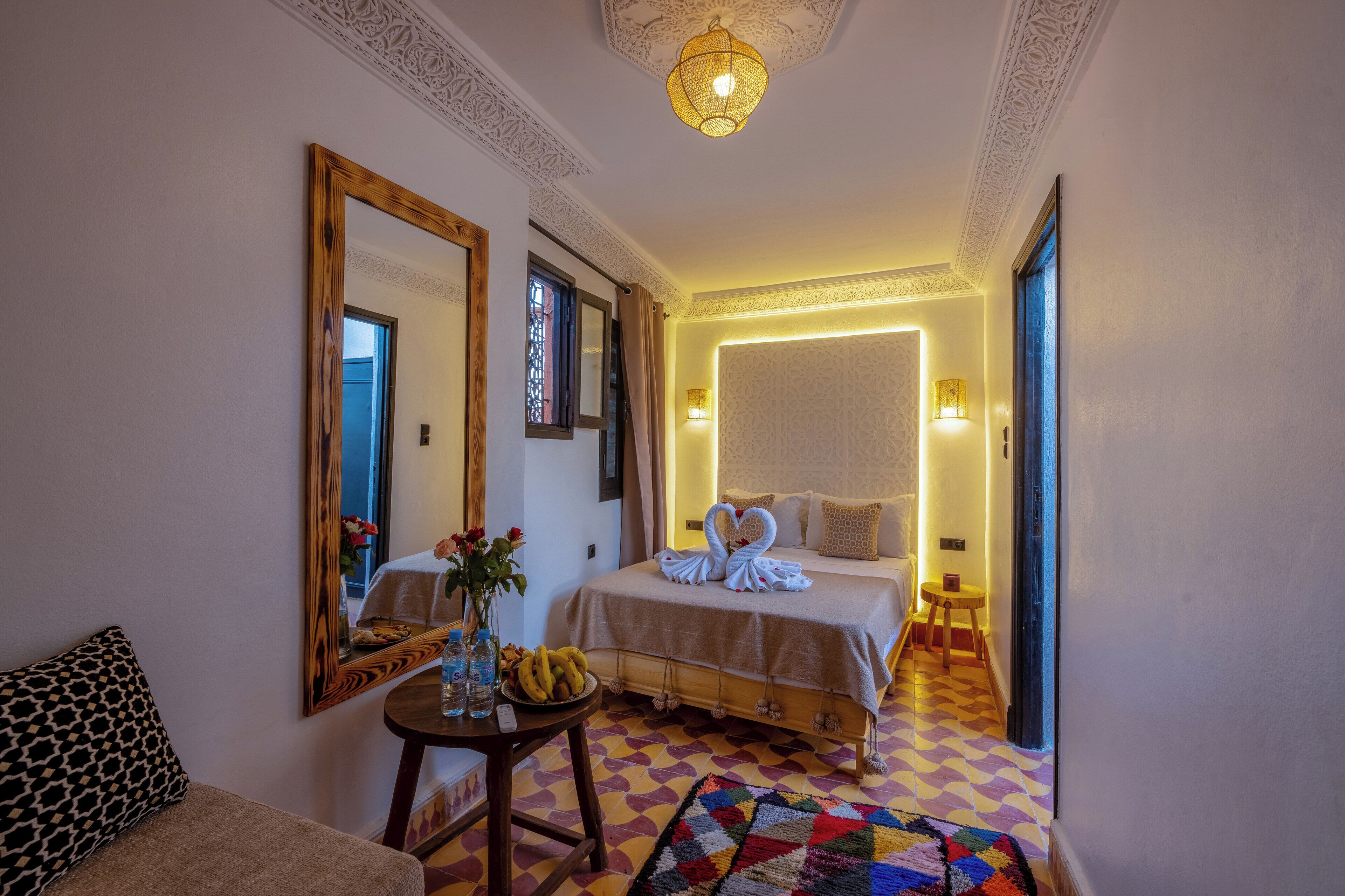 Una habitación de hotel con cama, un espejo, una mesita con un florero y bananas, y una alfombra colorida.