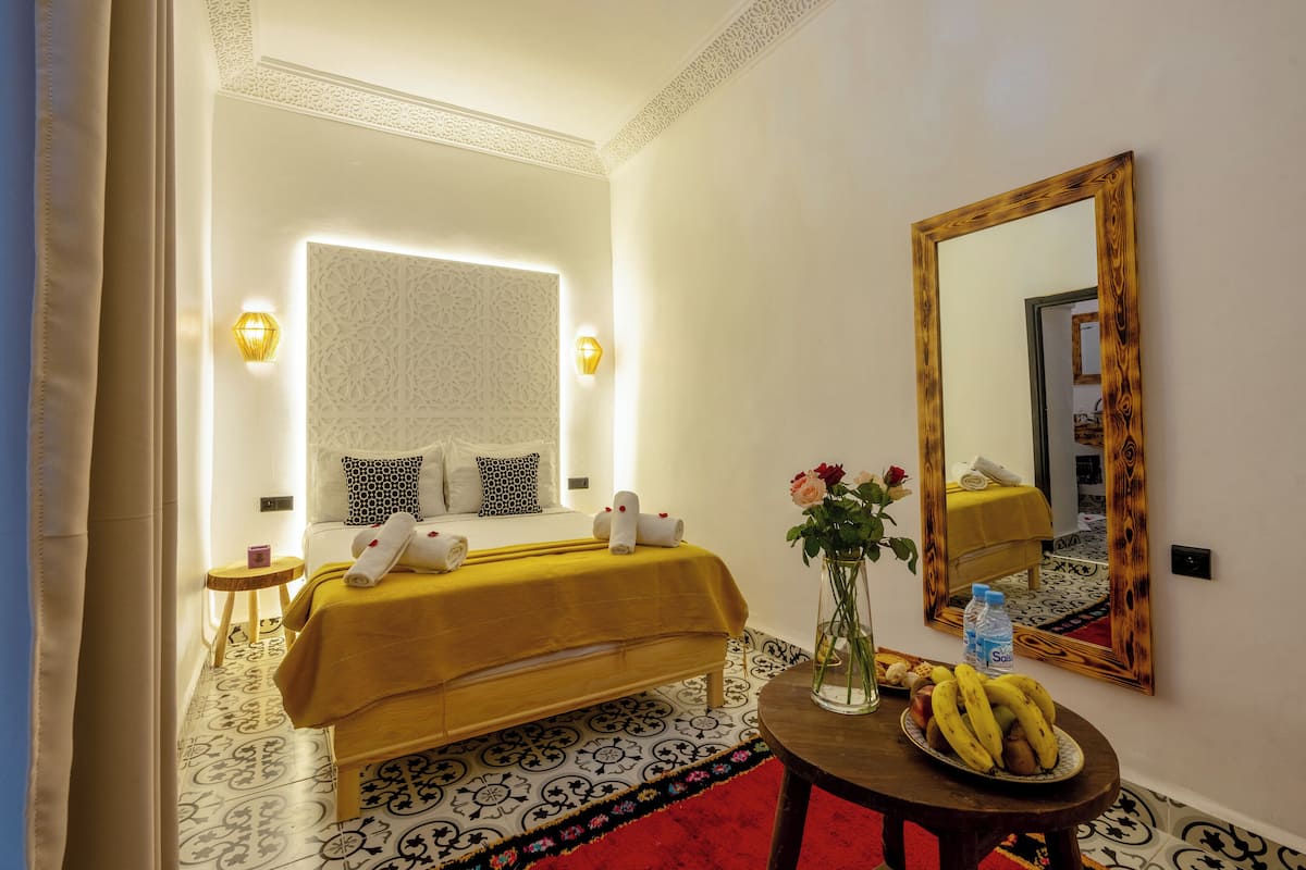 A hotel room with a bed, a round table with a fruit bowl, a vase with flowers, a mirror, and a patterned floor.