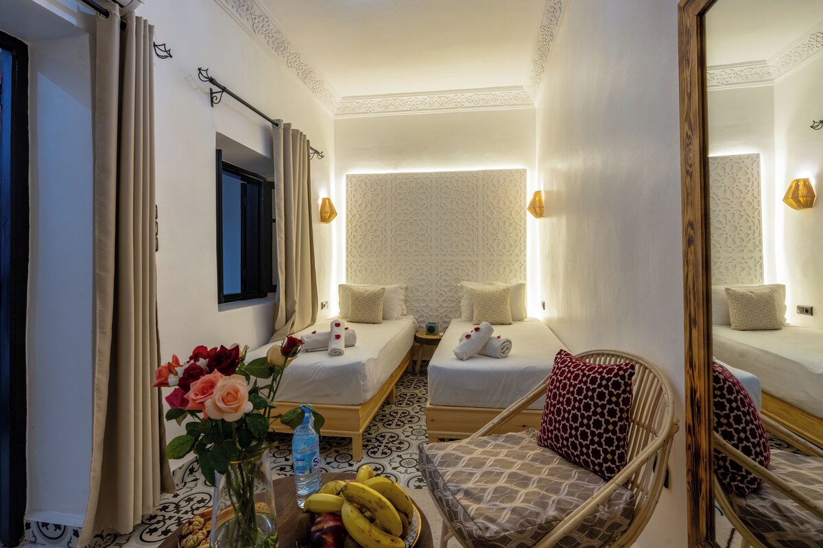 A hotel room with two beds, a small table with a vase of flowers, a chair, and a mirror.