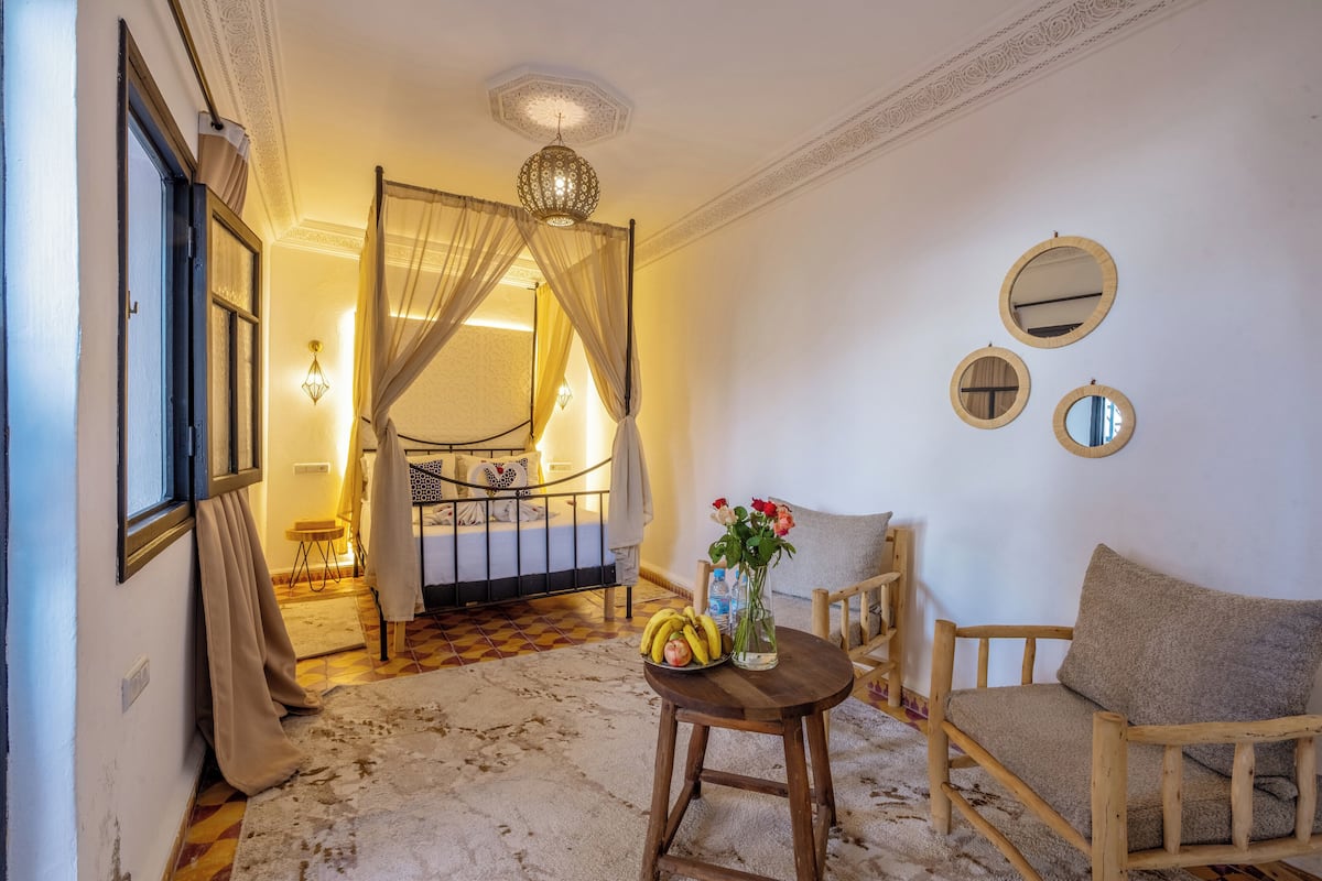 A cozy room with a bed, a wooden chair, a small table with a vase and bananas, and a decorative ceiling light.