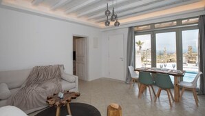 Een moderne woonkamer met een bank, een salontafel en een eethoek met een tafel en stoelen. Er is een groot raam met uitzicht op een zwembad en palmbomen.