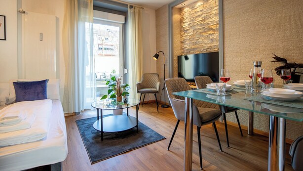 A modern hotel room with a bed, a glass dining table, chairs, a small side table with a plant, a TV, and a stone wall. - Special *Roger F. Rabbit 1* Apartment (Basel)
