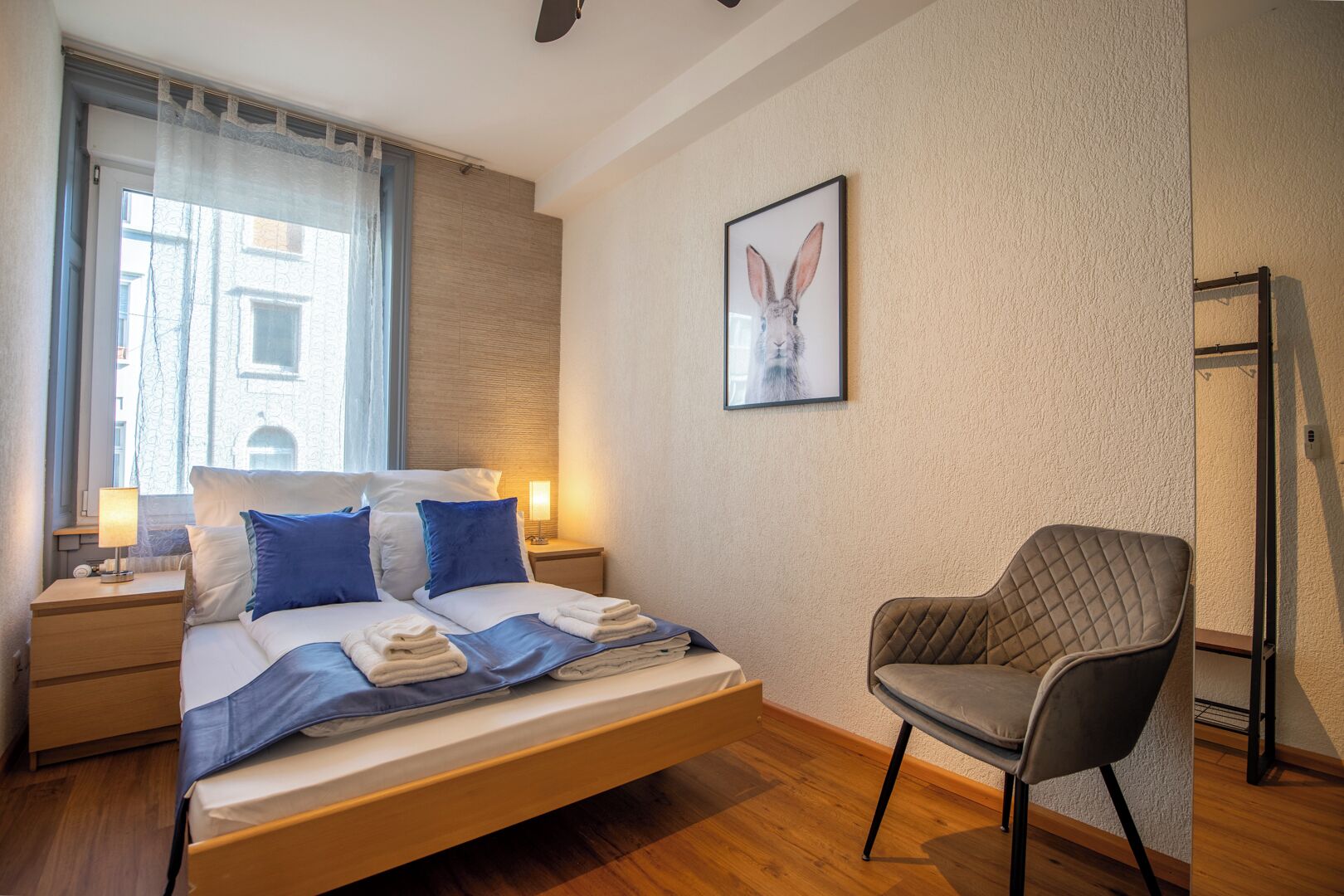 A hotel room with a bed, bedside tables, a chair, a window with curtains, a wall-mounted picture, and a door.
