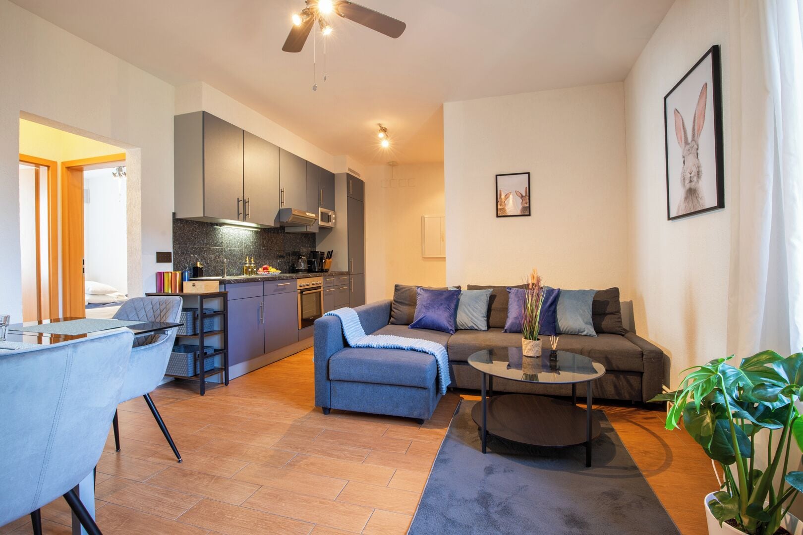 Una sala moderna con sofá, reposapiés y mesa de centro. Se observa un comedor con una mesa y sillas. Se puede ver la cocina con armarios y electrodomésticos.