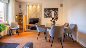 A modern living room with a flat-screen TV, a dining table with four chairs, a bookshelf, and a decorative wall panel. - Special *Roger F. Rabbit 2* Apartment (Basel)