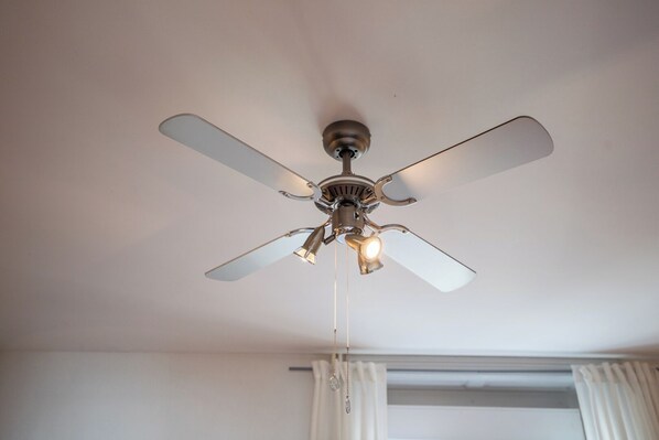 Un ventilateur de plafond à quatre pales et un luminaire dans une pièce aux murs blancs et dotée d’une fenêtre avec des rideaux.