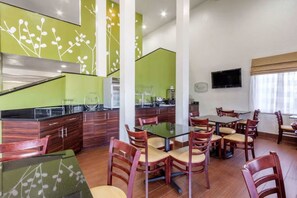 Un espace repas moderne avec une table en verre entourée de chaises en bois, un coin cuisine agrémenté de touches de vert et un téléviseur à écran plat fixé au mur.
