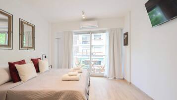 Una habitación de hotel con una cama grande, un televisor de pantalla plana, un balcón con mesa y sillas, y vistas a un edificio con balcones.