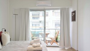 Habitación de hotel con cama, mesita de noche, lámpara, ventana con cortinas y vistas al balcón exterior.