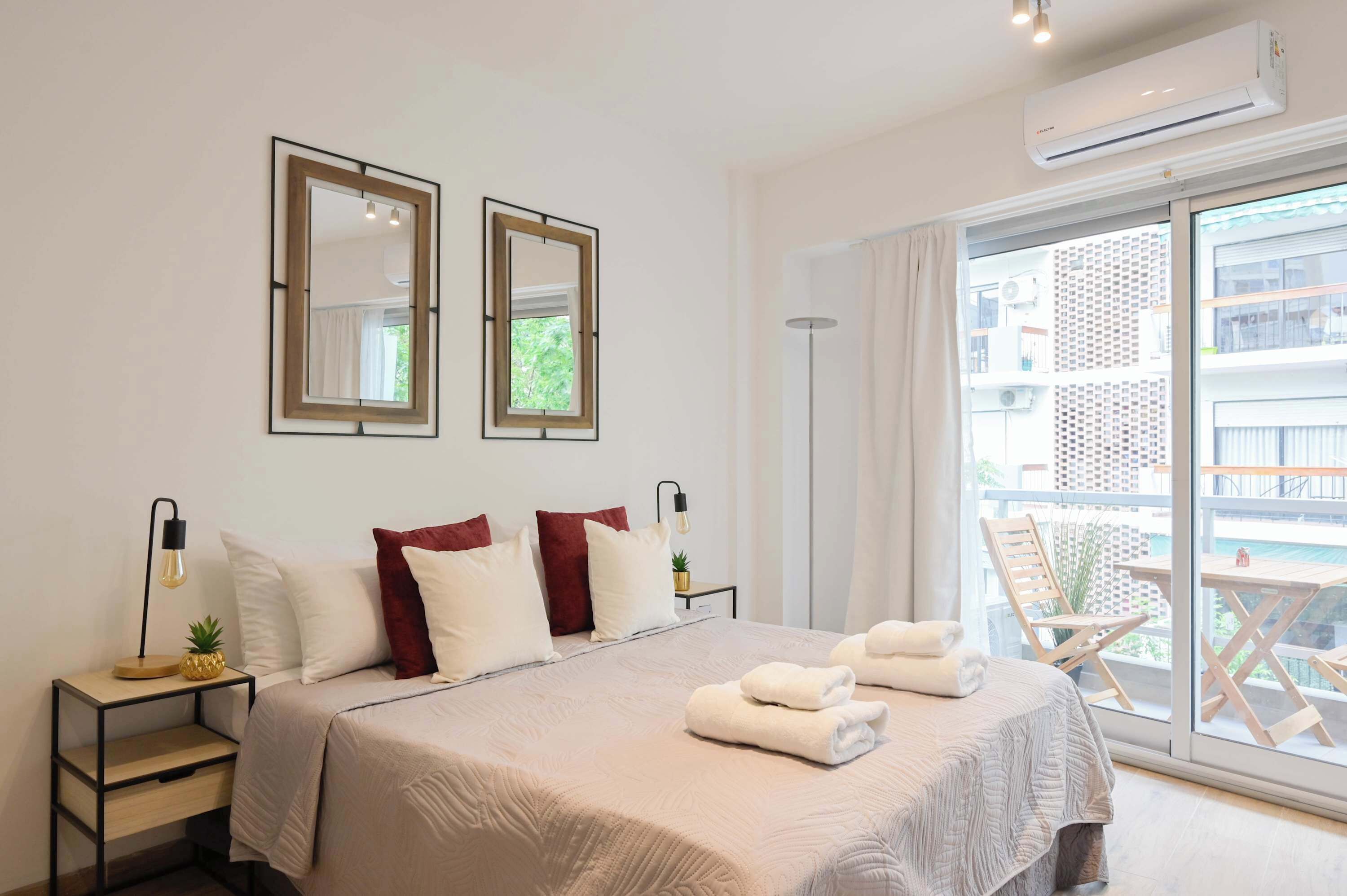 Habitación de hotel con una cama grande, dos mesitas de noche con lámparas, una ventana con vistas a edificios y un equipo de aire acondicionado.