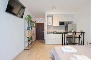 A compact hotel room with a kitchenette, a bed with towels, a bookshelf, and a door leading to another room.