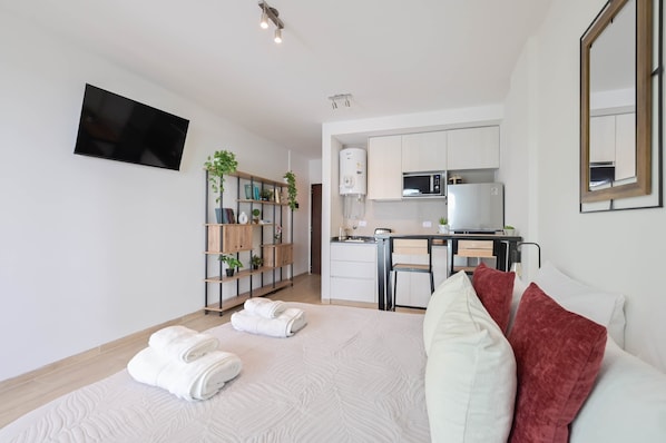 A modern hotel room with a large bed, a flat-screen TV mounted on the wall, a kitchenette with a microwave and refrigerator, and a small desk area.