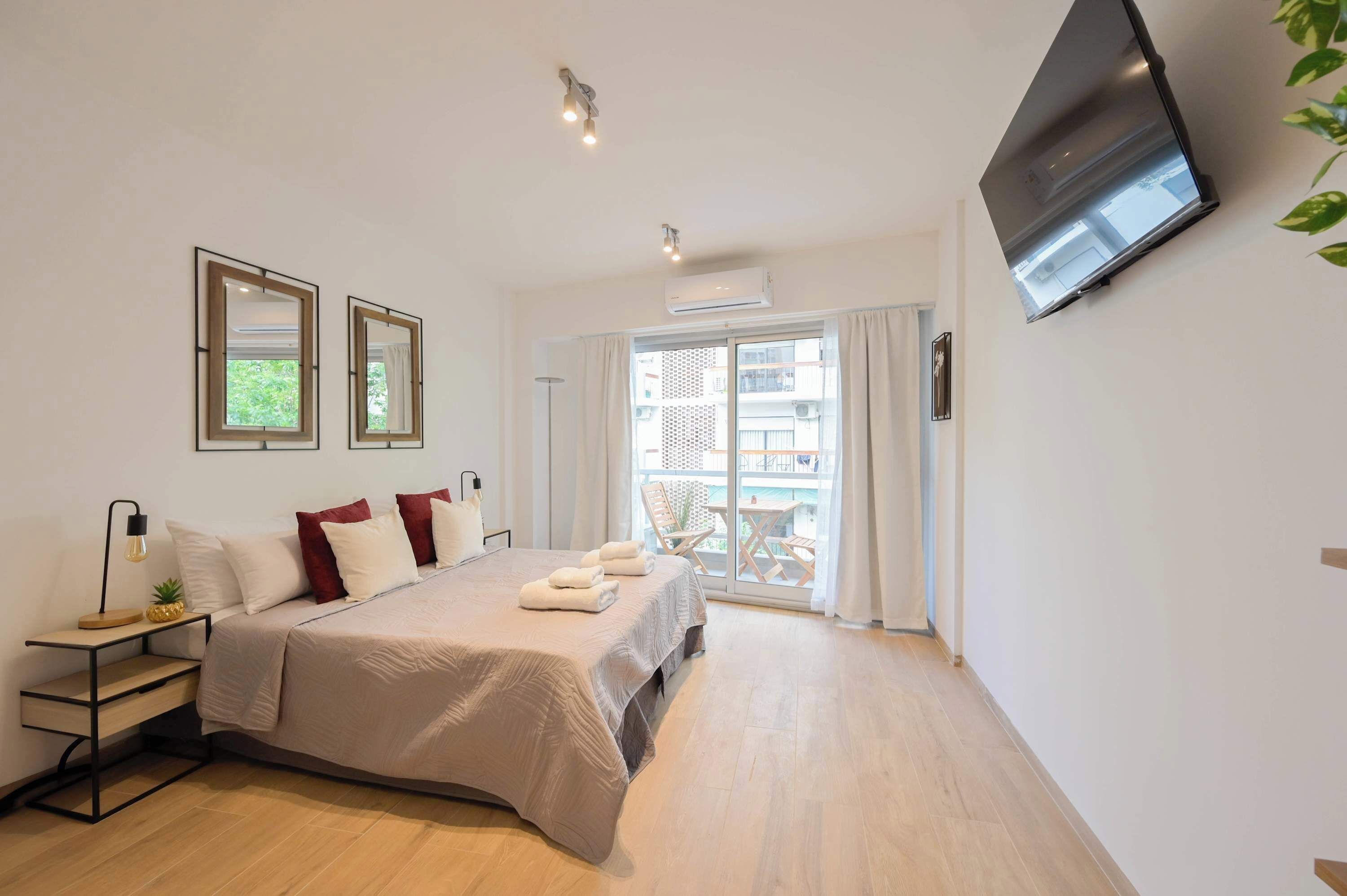 A modern hotel room with a bed, bedside table, lamp, air conditioning unit, and a TV mounted on the wall.