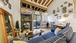 Una sala de estar acogedora con un sofá, una mesa de centro y una chimenea. Cuenta con un comedor con una mesa y sillas. La habitación tiene vigas de madera en el techo y un ventanal grande.