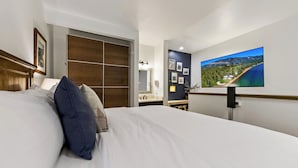 Una habitación de hotel con una cama grande, un televisor montado en la pared, un armario y un baño con espejo y lavamanos.