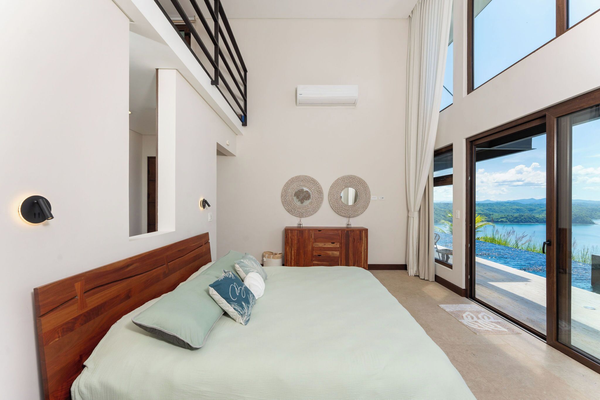 A bedroom with a large bed, a wooden headboard, a nightstand, two wall-mounted mirrors, and a view of a lake through a sliding glass door.
