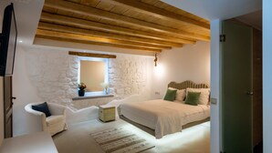 Une chambre avec un mur en pierre, un plafond en bois, un lit avec des coussins verts, une chaise blanche et une petite table.