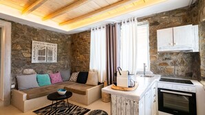 Un salon chaleureux avec un mur en pierre, un canapé agrémenté de coussins à motifs, une petite table avec un saladier, une cuisine équipée d’un évier et d’un four, et une fenêtre avec des rideaux.