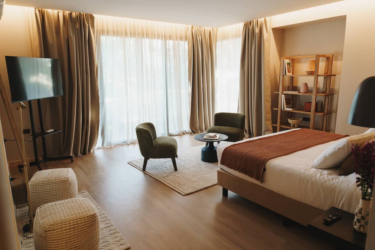 A modern hotel room with a large bed, two armchairs, a coffee table, a TV, and a bookshelf.